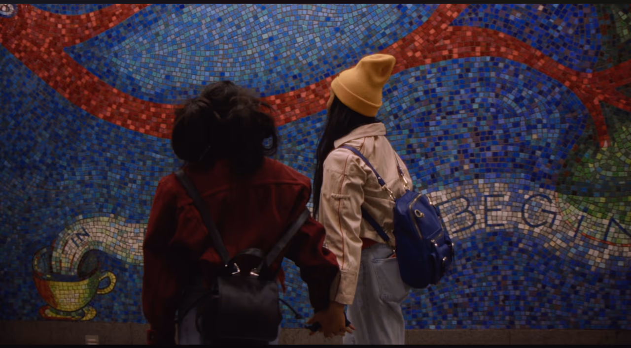 Two women hold hands with their backs to the camera, facing a blue mosaic wall with a red ribbon and a teacup