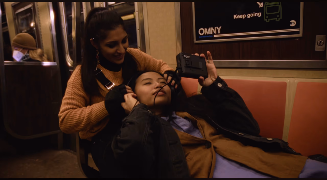 On a subway car, a woman in an orange sweater puts headphones on a friend lying across her lap holding a camera