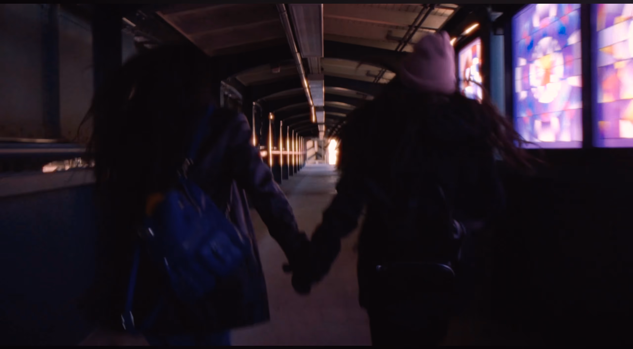 Two people walk hand in hand away from the camera down a covered walkway lit by stained-glass panels