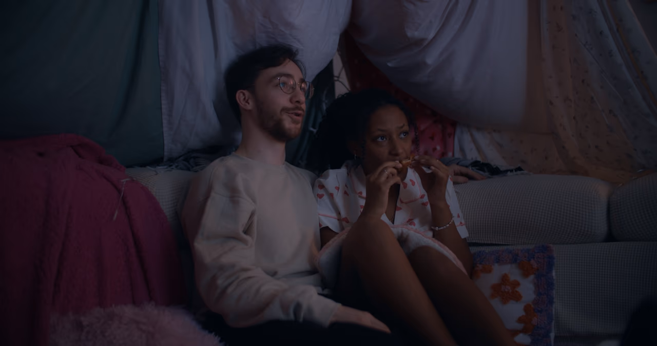 A man in glasses and a woman eating a snack sit side by side on a sofa inside a bedsheet fort