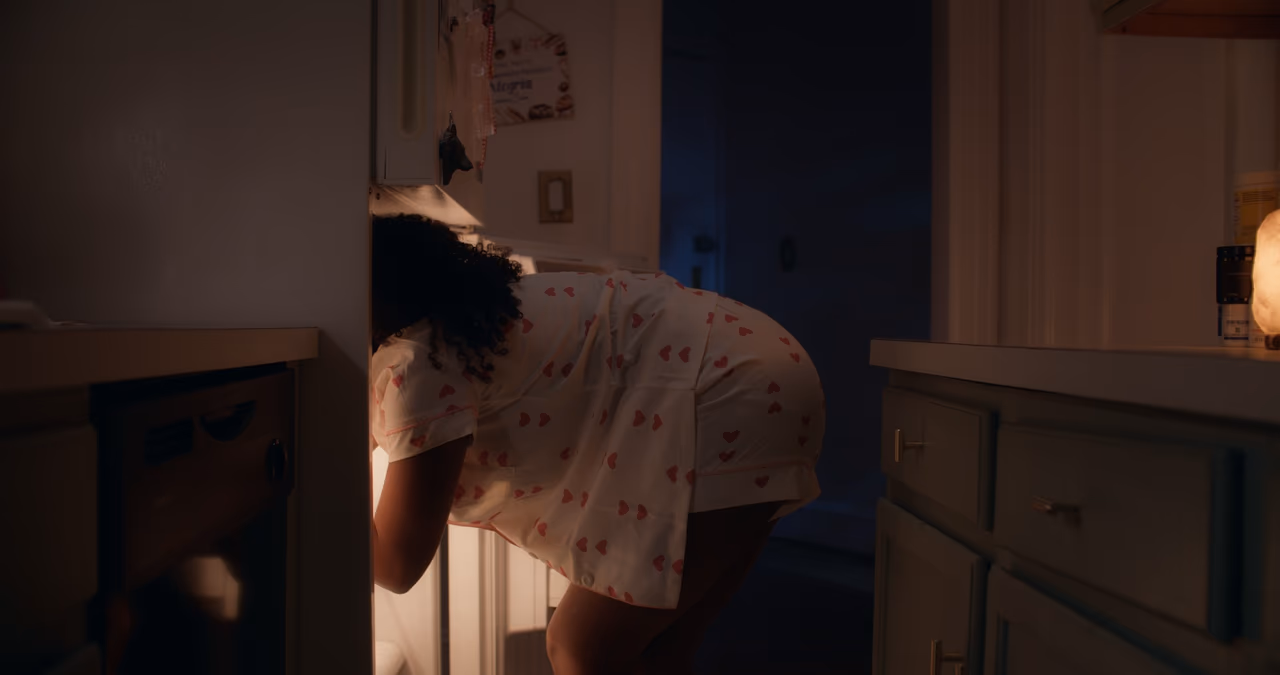 A woman in heart-print pyjamas leans into the lit refrigerator of a dark kitchen at night