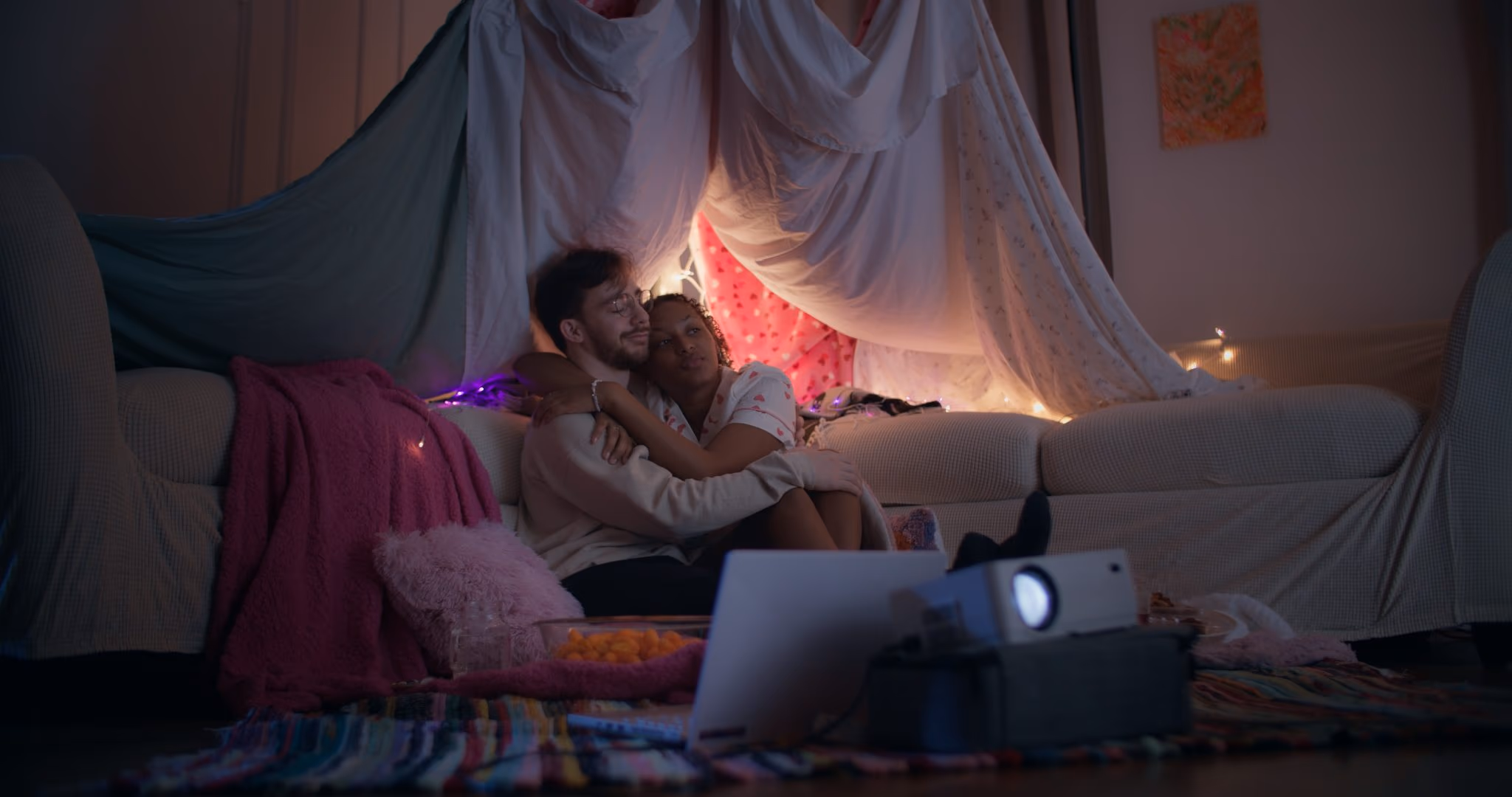 A couple hugs on the floor of a bedsheet fort strung with fairy lights, a projector and laptop in front of them