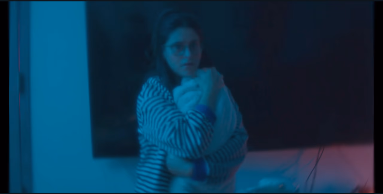 A woman in a striped top and glasses clutches a bundled blanket to her chest in a blue-lit room