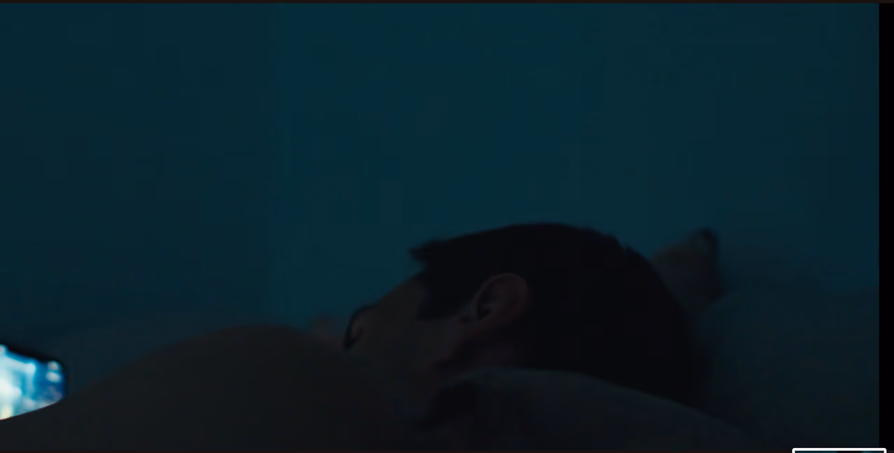 The back of a man's head on a pillow in a dark teal bedroom, a phone screen glowing at the edge of the frame