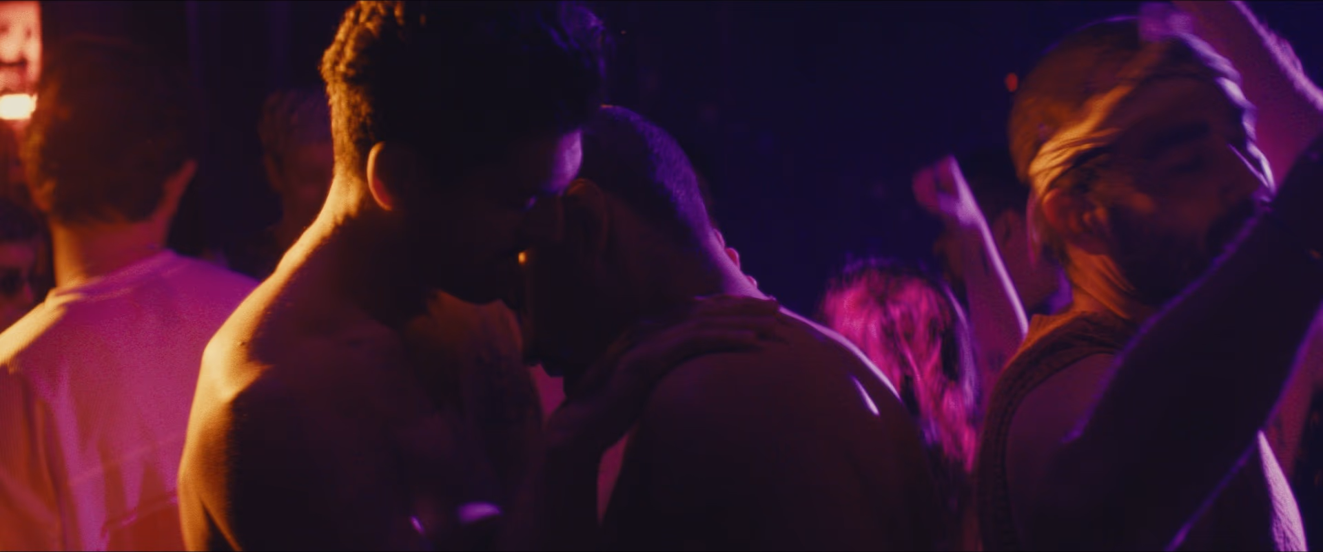Two men lean close together in a crowd on a dance floor washed in pink and violet light