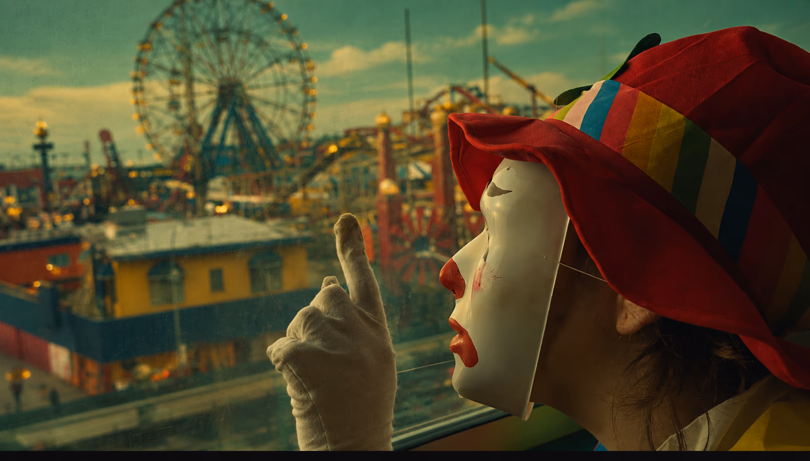 A clown in a white mask, gloves and a red rainbow-striped hat looks out a window at a Ferris wheel and rides